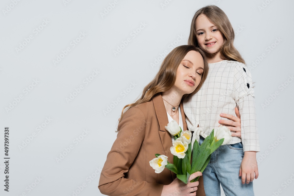 happy girl looking at camera near mother with tulips isolated on grey.