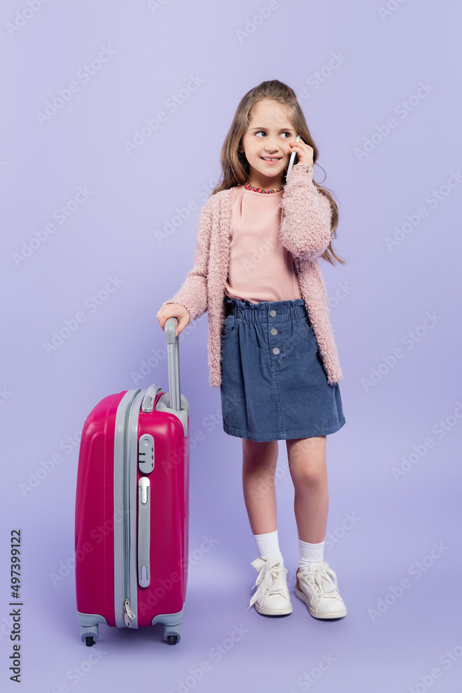 full length of happy girl talking on smartphone near pink baggage on purple. full length of happy girl talking on smartphone near pink baggage on purple.