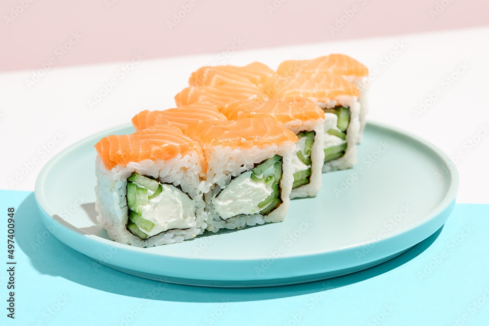 Classic sushi roll with salmon outside, cheese and cucumber inside. Philadelphia maki in minimal style. Modern japanese menu concept. Salmon maki roll on ceramic plate on coloured background. Classic sushi roll with salmon outside, cheese and cucumber inside. Philadelphia maki in minimal style. Modern japanese menu concept. Salmon maki roll on ceramic plate on coloured background.