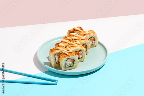 Tempura maki roll on ceramic plate with chopsticks. Hot sushi with salmon, crab and cucumber inside, spicy mayo topped. Modern japanese menu concept. Maki sushi on coloured background.