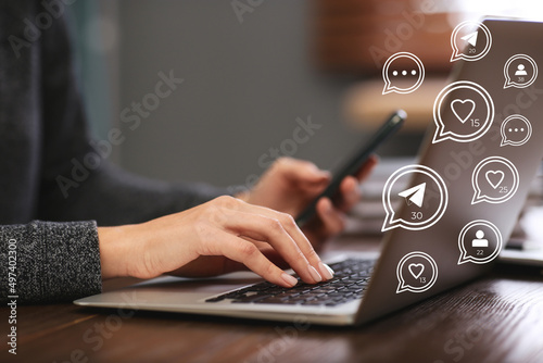 Different virtual icons and young woman using laptop and smartphone at table indoors, closeup. SMM (Social media marketing) concept photo