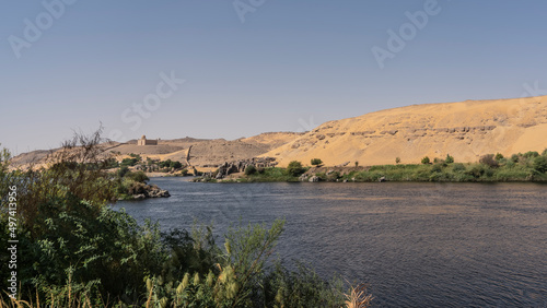 Fototapeta Naklejka Na Ścianę i Meble -  Green vegetation grows on the banks of the Nile River. Ripples on the blue water. High sand dunes against a clear azure sky. Egypt. Aswan