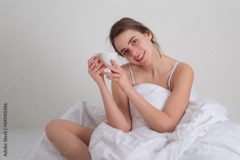 Morning leisure. Happy young woman with cup of coffee or tea in bed at home bedroom. Portrait of beautiful woman in white pajamas drinking coffee. Morning leisure. Happy young woman with cup of coffee or tea in bed at home bedroom. Portrait of beautiful woman in white pajamas drinking coffee.