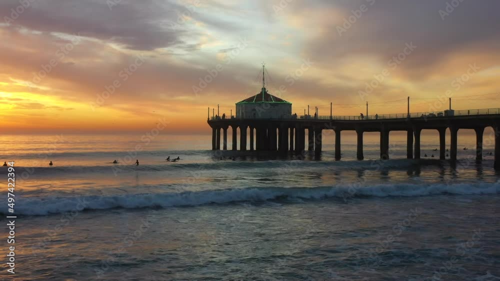 People Surfing Under the Manhattan Beach Pier at Sunset with a Lit Up Roundhouse Aquarium Roof in California USA - Forward Panning Wide Shot