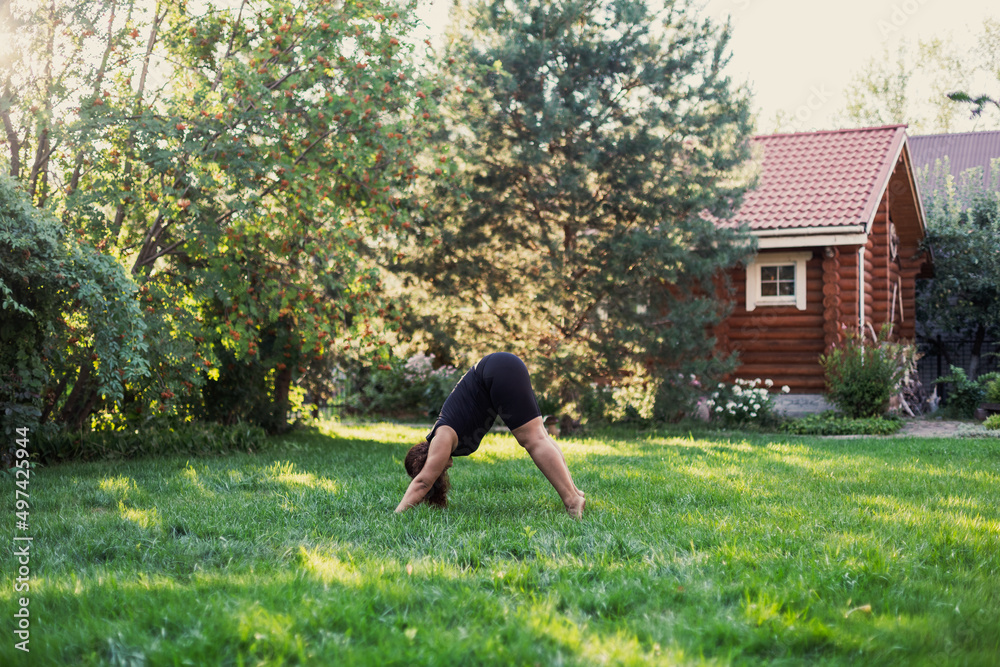 Over-weighted female doing yoga standing in triangle position on backyard of cottage with wooden house and trees in background. Body positive. Equality. 