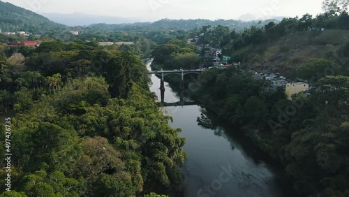 Several vehicles drive left-handed over the old high Peradeniya Bridge over the reflective Mahaweli Ganga River on a sunny day among the green nature of Sri Lanka. Drone dolley shot to close up photo