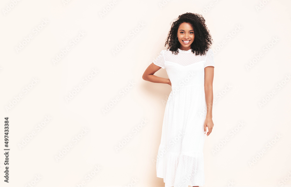 Beautiful black woman with afro curls hairstyle. Smiling model dressed in white summer dress. Sexy carefree female posing near wall in studio. Tanned and cheerful. Isolated Beautiful black woman with afro curls hairstyle. Smiling model dressed in white summer dress. Sexy carefree female posing near wall in studio. Tanned and cheerful. Isolated