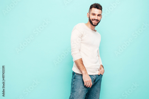 Portrait of handsome smiling stylish hipster lambersexual model.Man dressed in white sweater and jeans. Fashion male isolated on blue wall in studio