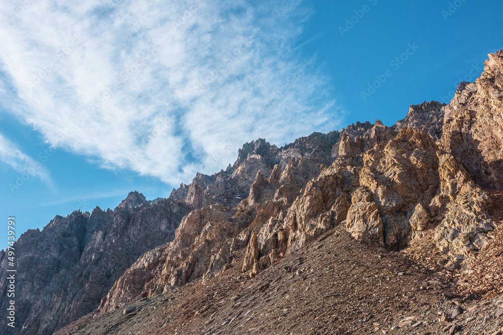 Scenic mountain landscape with sharp rocks under cirrus clouds in sunny day. Colorful scenery with gold sunlit sharp rocky mountains. High rocky mountains in golden sunlight under spindrift clouds.