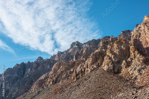 Scenic mountain landscape with sharp rocks under cirrus clouds in sunny day. Colorful scenery with gold sunlit sharp rocky mountains. High rocky mountains in golden sunlight under spindrift clouds.
