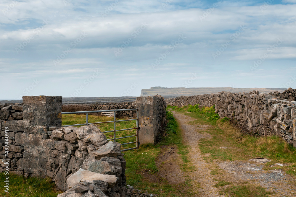 Small road between stone walls and entrance to a agriculture field. Inishmore, Aran island, Ireland. Blue cloudy sky. Popular tourist area. Dún Aonghasa in the background