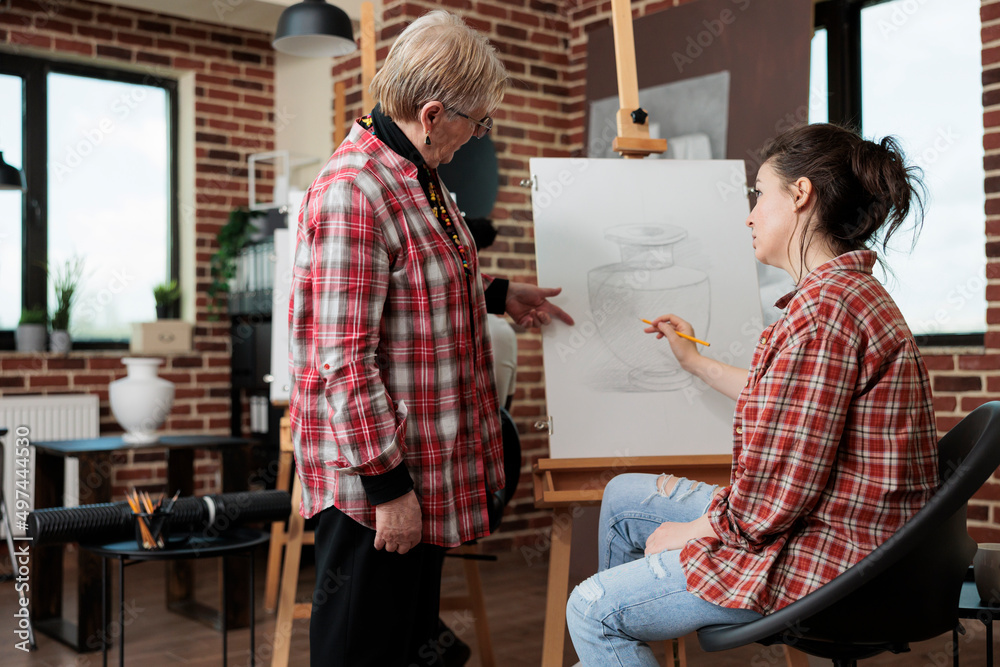 Fototapeta premium Senior art teacher showing graphic technique to young student during creativity lesson in school studio. Painter woman drawing vase sketch using sketching pencil working at personal growth