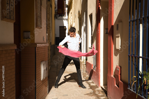 Young Spanish man in white shirt and black pants and dance shoes, dancing flamenco with a red handkerchief with white polka dots in a very narrow street. Concept art, dance, culture, tradition.