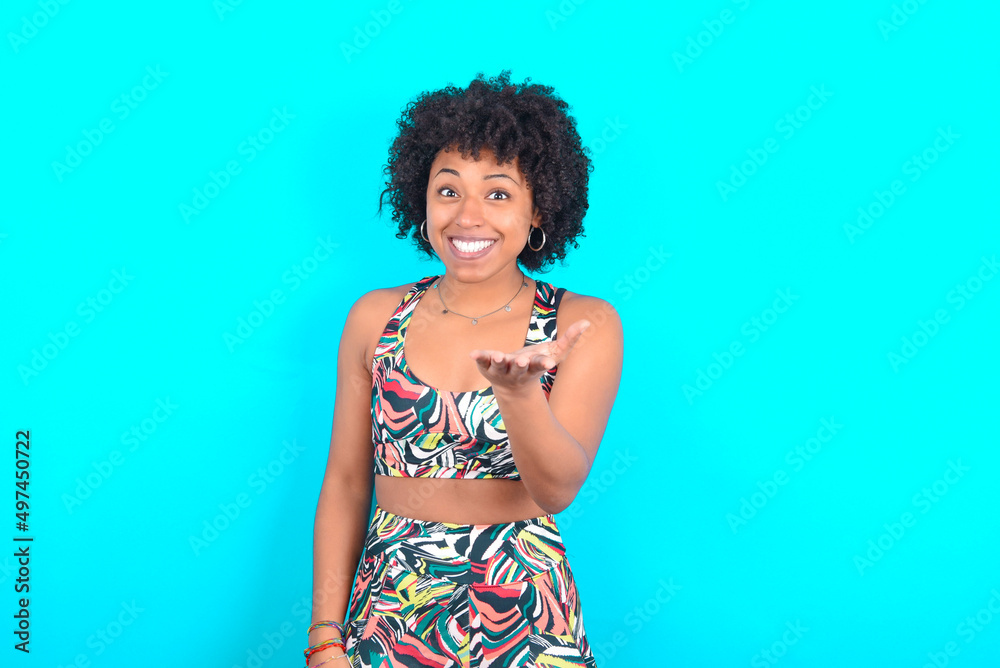young woman with afro hairstyle in sportswear against blue background smiling friendly offering something with open hand or handshake as greeting and welcoming. Successful business.