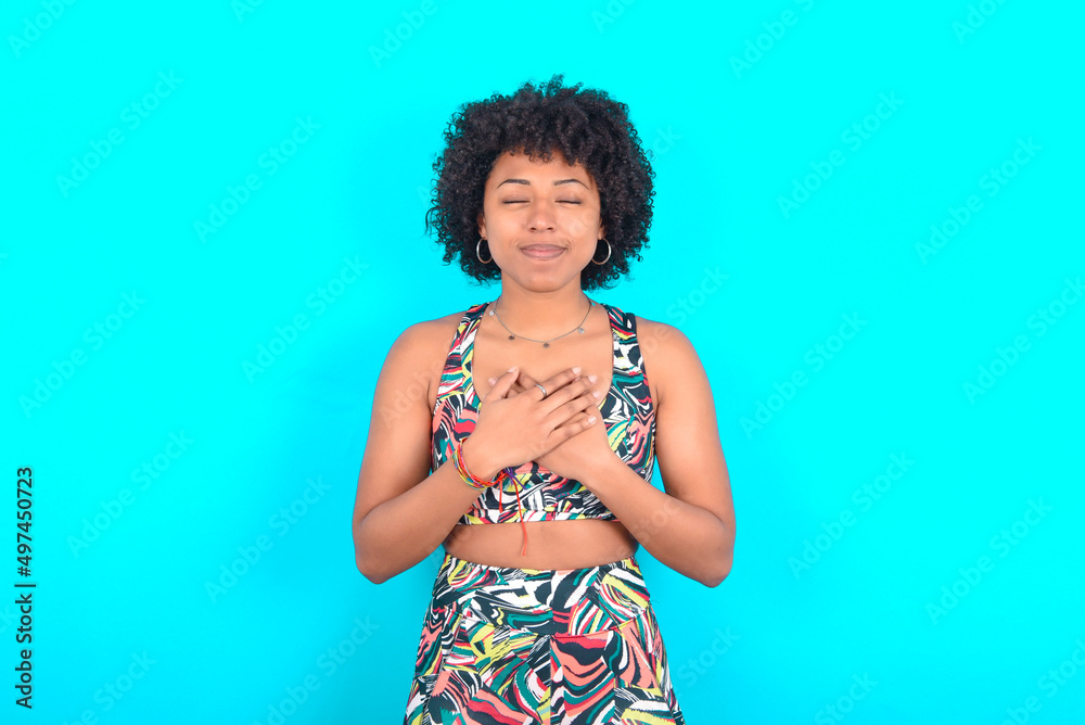 young woman with afro hairstyle in sportswear  closes eyes and keeps hands on chest near heart, expresses sincere emotions, being kind hearted and honest. Body language and real feelings concept.