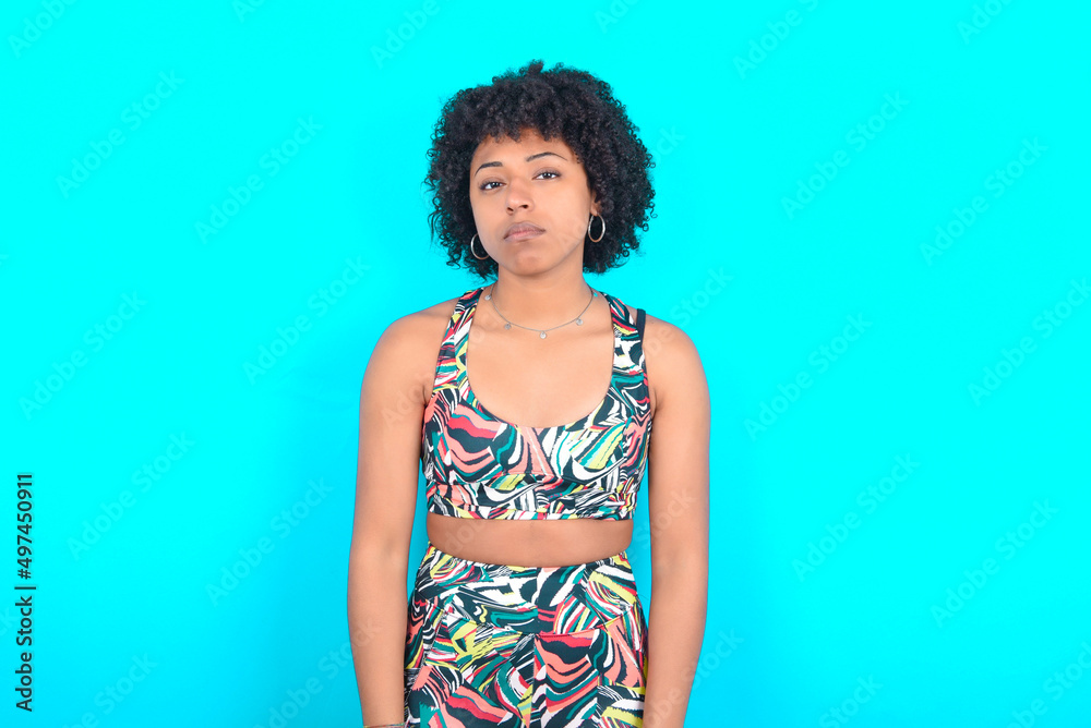 Gloomy, bored young woman with afro hairstyle in sportswear against blue background frowns face looking up, being upset with so much talking hands down, feels tired and wants to leave. Gloomy, bored young woman with afro hairstyle in sportswear against blue background frowns face looking up, being upset with so much talking hands down, feels tired and wants to leave.