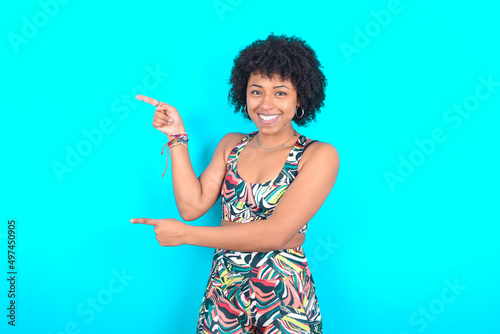 young woman with afro hairstyle in sportswear against blue background points aside with  surprised expression with mouth opened, shows something amazing. Advertisement concept.