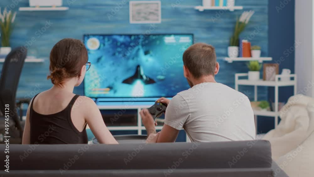 Girlfriend eating popcorn and supporting boyfriend gamer playing console video game first person shooter on tv while sitting on sofa. Couple on couch enjoying gaming simulation in modern living room. Girlfriend eating popcorn and supporting boyfriend gamer playing console video game first person shooter on tv while sitting on sofa. Couple on couch enjoying gaming simulation in modern living room.
