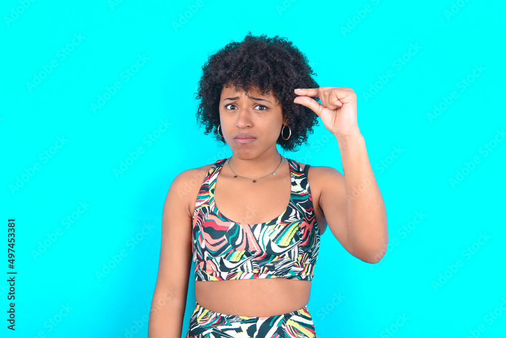 Upset young woman with afro hairstyle in sportswear against blue background shapes little gesture with hand demonstrates something very tiny small size. Not very much Upset young woman with afro hairstyle in sportswear against blue background shapes little gesture with hand demonstrates something very tiny small size. Not very much