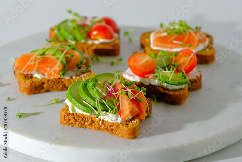 closeup healthy toast with salmon, trout, cherry tomatoes, avocado and microgreens on white marble board