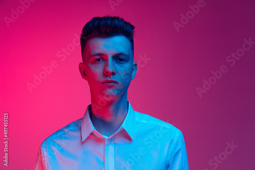 Half-length portrait of young handsome man in white shirt posing isolated over magenta color studio background in blue neon light