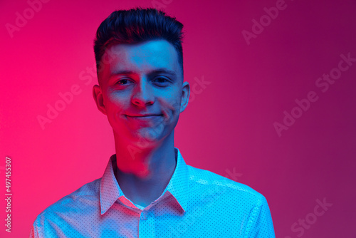 Young happy man in white shirt smiling isolated over magenta color studio background in blue neon light. Concept of positive emotions