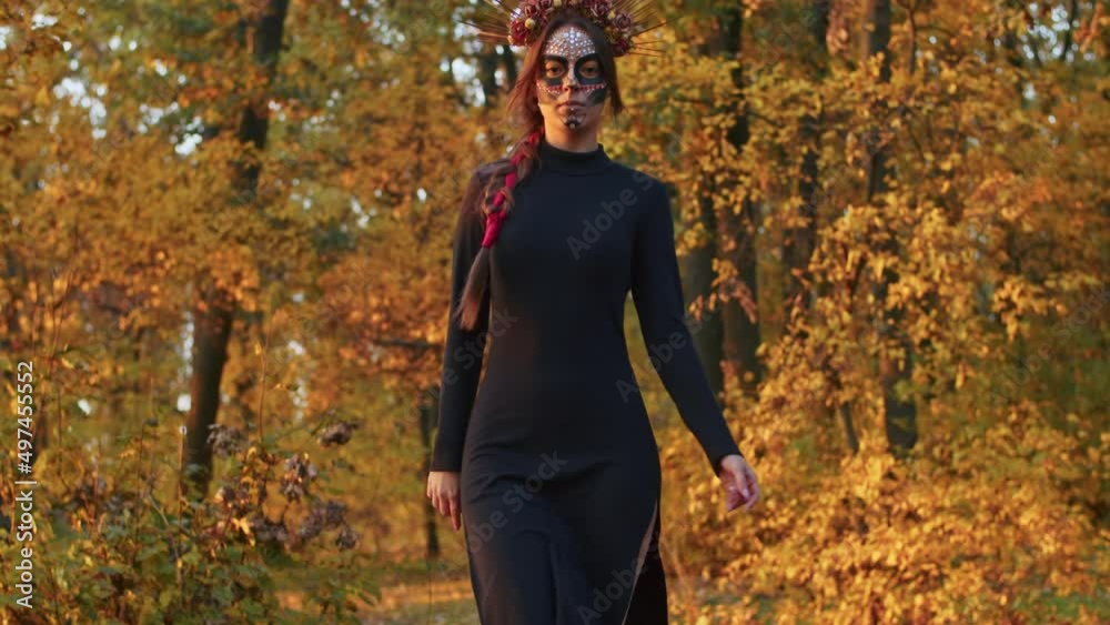 A young woman with Santa Muerte make-up dressed in a black dress of death walks against the backdrop of autumn leaves in the forest during sunset. Day of the Dead or Halloween concept. A young woman with Santa Muerte make-up dressed in a black dress of death walks against the backdrop of autumn leaves in the forest during sunset. Day of the Dead or Halloween concept.