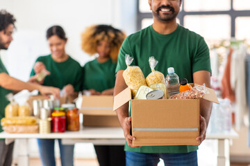 charity, donation and volunteering concept - happy smiling male volunteer with food in box and international group of people at distribution or refugee assistance center