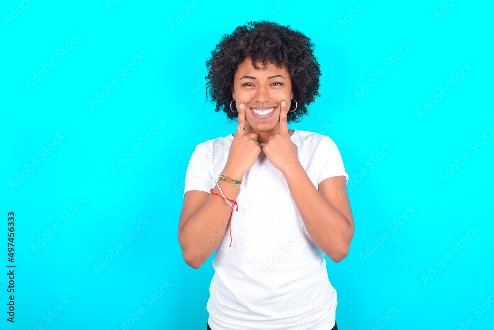 Happy young woman with afro hairstyle wearing white T-shirt against blue wall with toothy smile, keeps index fingers near mouth, fingers pointing and forcing cheerful smile Happy young woman with afro hairstyle wearing white T-shirt against blue wall with toothy smile, keeps index fingers near mouth, fingers pointing and forcing cheerful smile