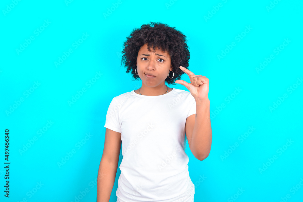Displeased young woman with afro hairstyle wearing white T-shirt against blue wall shapes little hand sign demonstrates something not very big. Body language concept.