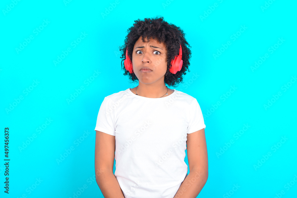 Serious displeased young woman with afro hairstyle wearing white T-shirt against blue wall looks puzzled at camera being angry wears stereo headphones listens music while walking at street Serious displeased young woman with afro hairstyle wearing white T-shirt against blue wall looks puzzled at camera being angry wears stereo headphones listens music while walking at street