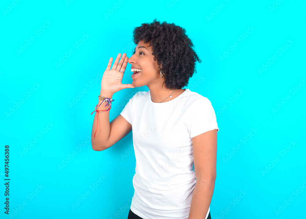 young woman with afro hairstyle wearing white T-shirt against blue wall look empty space holding hand near her face and screaming or calling someone. young woman with afro hairstyle wearing white T-shirt against blue wall look empty space holding hand near her face and screaming or calling someone.