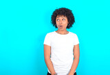 Shocked young woman with afro hairstyle wearing white T-shirt against blue wall look empty space with open mouth screaming: Oh My God! I can't believe this.