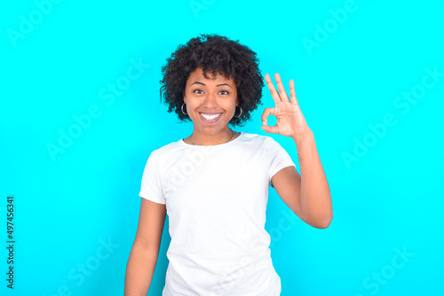 young woman with afro hairstyle wearing white T-shirt against blue wall hold hand arm okey symbol toothy approve advising novelty news