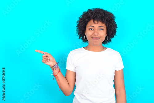 young woman with afro hairstyle wearing white T-shirt against blue wall points to side on blank space demonstrates advertisement. People and promotion concept