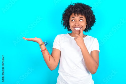 Positive young woman with afro hairstyle wearing white T-shirt against blue wall advert promo touches teeth with finger.