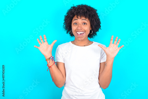 Delighted positive young woman with afro hairstyle wearing white T-shirt against blue wall opens mouth and arms palms up after having great result