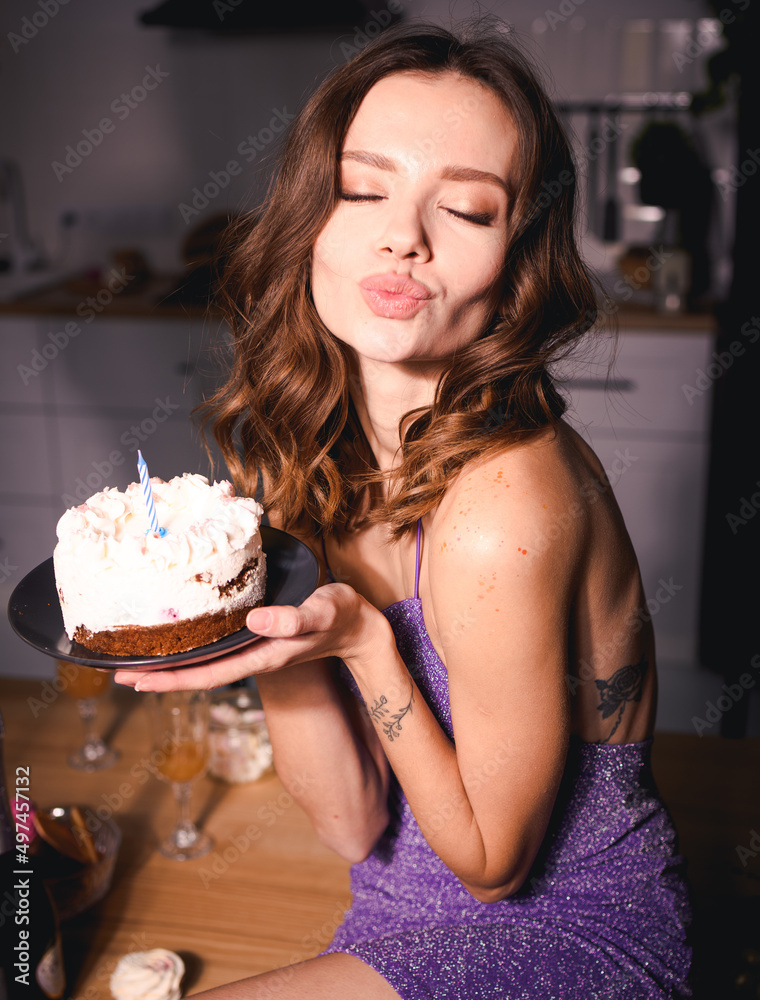 Young woman in dress holding birthday cake with candle and blowing air kiss in kitchen Young woman in dress holding birthday cake with candle and blowing air kiss in kitchen