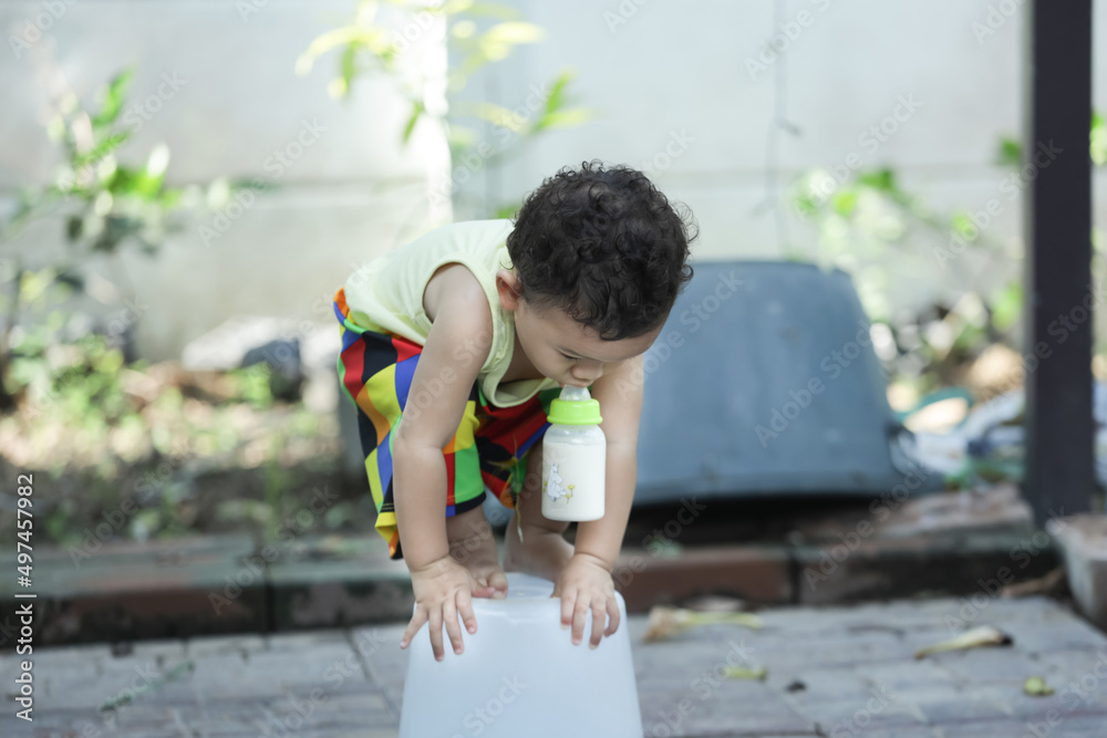 A child is feeding from a bottle and is standing on a plastic bucket.