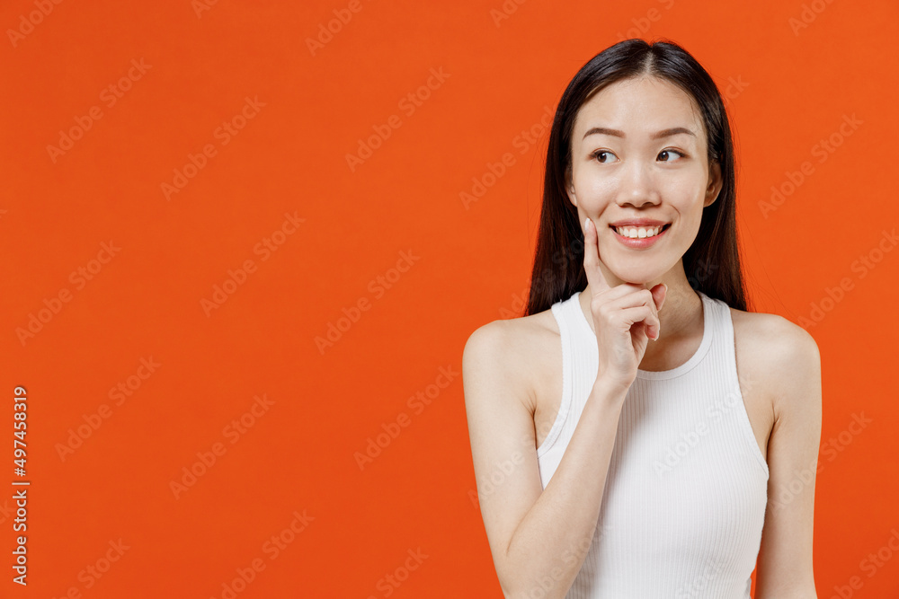 Fun young woman of Asian ethnicity 20s years old in white tank top look around think dream put hand prop up on chin lost in thought and conjectures isolated on plain orange background studio portrait. Fun young woman of Asian ethnicity 20s years old in white tank top look around think dream put hand prop up on chin lost in thought and conjectures isolated on plain orange background studio portrait.