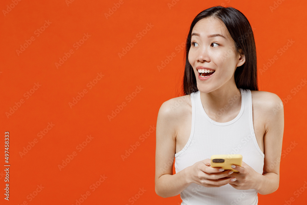 Surprised young woman of Asian ethnicity 20s years old in white tank top hold in hand use mobile cell phone typing browsing send sms looking aside isolated on plain orange background studio portrait. Surprised young woman of Asian ethnicity 20s years old in white tank top hold in hand use mobile cell phone typing browsing send sms looking aside isolated on plain orange background studio portrait.