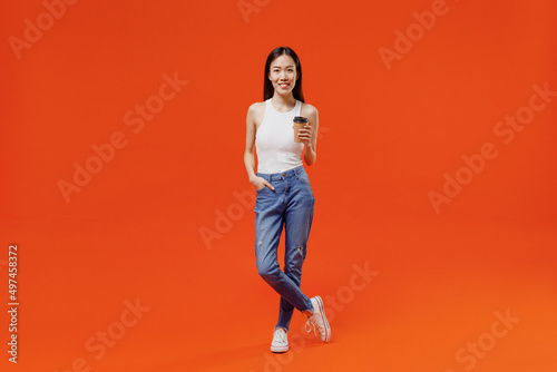 Full size smiling excited young woman of Asian ethnicity 20s years old in white tank top hold takeaway delivery craft paper brown cup coffee to go isolated on plain orange background studio portrait.