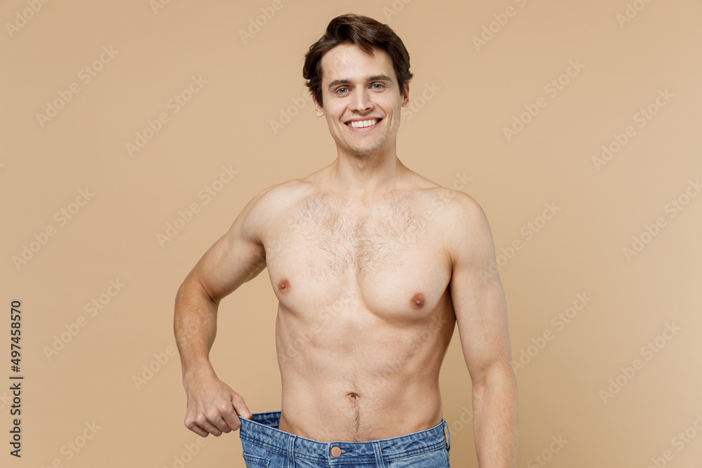 Satisfied shirtless young man 20s with naked torso showing loose pants isolated on pastel pastel beige color background Body care healthcare fitness sport bodybuilding diet loosing weight concept.