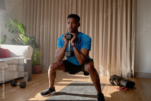 Young African American male exercising from home concentrating while squatting with a dumbbell in living room, workout at home