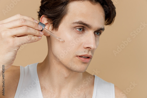 Closeup young man 20s perfect skin in undershirt hold apply bottle face body oil, moisturizing serum isolated on pastel pastel beige background studio Skin care healthcare cosmetic procedures concept