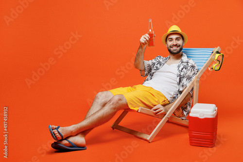 Full size young happy cool tourist man in beach shirt hat hold beer bottle lie on deckchair near fridge isolated on plain orange background studio portrait. Summer vacation sea rest sun tan concept.