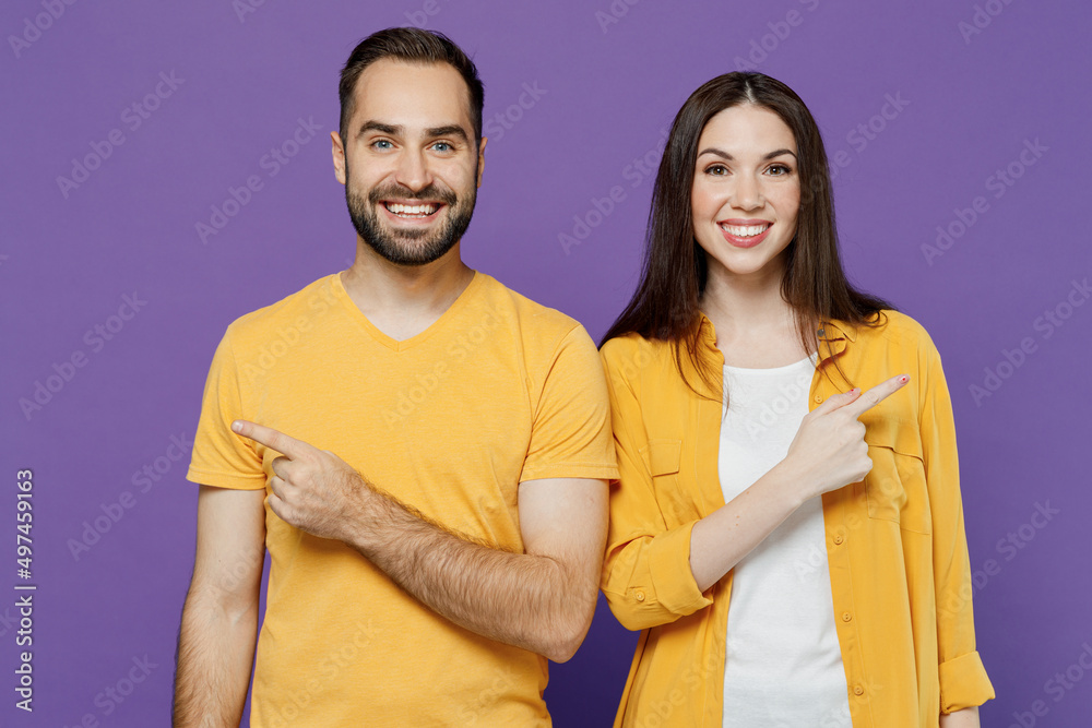 Obraz premium Young smiling happy couple two friends family man woman together in yellow casual clothes looking camera point index finger aside on workspace area isolated on plain violet background studio portrait.