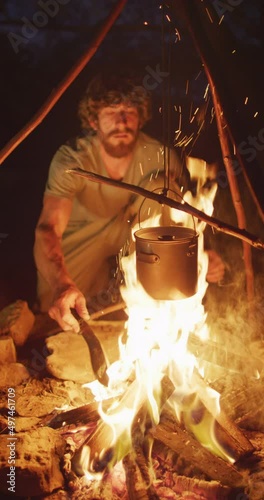 Vertical video of bearded caucasian male survivalist cooking over campfire in wilderness photo