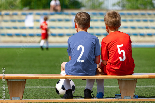 Two boys' sports friends sitting on a wooden bench and watching a soccer match. School kids in football uniforms. Happy children playing sports outdoor. Friendship between two opposite sports teams © matimix