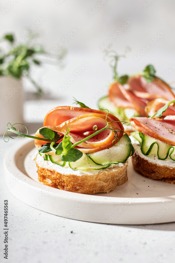 Three Bruschettas with baguette, bacon or meat, cream cheese, micro-greenery, fresh cucumber and sprouts, in composition on white plate on white textured background Three Bruschettas with baguette, bacon or meat, cream cheese, micro-greenery, fresh cucumber and sprouts, in composition on white plate on white textured background